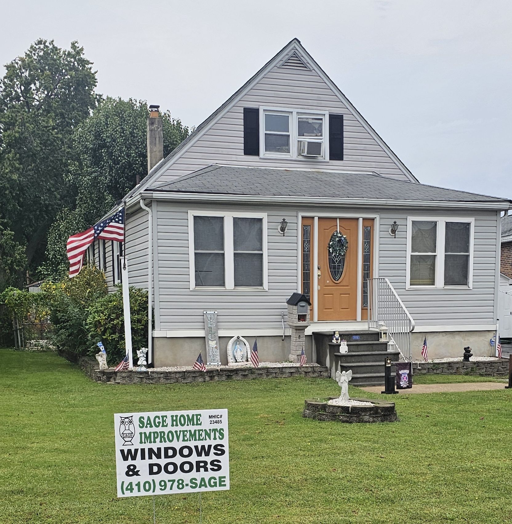 Gray Cape Cod–style home with white-trimmed windows, oak front door, and Sage Home Improvements yard sign