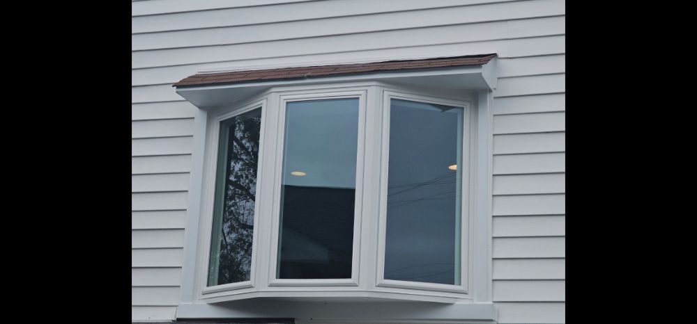 White three-pane bay window with brown shingled roof cap on a home with white horizontal siding